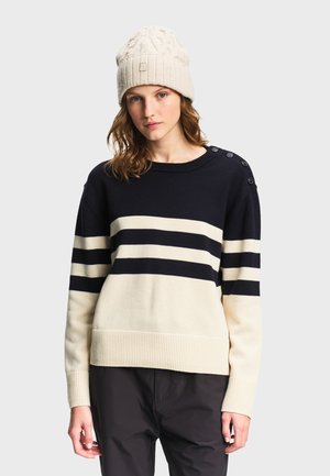 Donna che indossa un berretto di lana color crema e un maglione a righe nere e crema con bottoni sulla spalla, in piedi davanti a uno sfondo semplice.