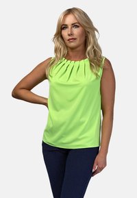 Top bez rękawów w jasnym neonowym zielonym kolorze, z plisowanym wzorem przy dekolcie. Gładki materiał o regularnym kroju, zestawiony z granatowymi spodniami w groszki.
