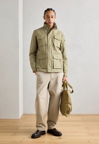 Jachetă utilitară verde măsliniu cu multiple buzunare frontale, purtată cu pantaloni cargo bej și cizme groase maro închis, ținând o geantă verde măsliniu.