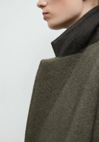 Abrigo de lana verde olivo con un cuello alto. Presenta un acabado texturizado y suave, con un acento de color oscuro en el cuello. Primer plano de la zona del hombro.