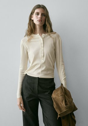 Frau mit langen Haaren, die ein beigefarbenes, langärmliges Oberteil mit Knöpfen und dunkle Hosen trägt, hält eine braune Wildlederjacke in der linken Hand.