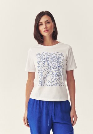 Kobieta w białej koszuli z krótkim rękawem z abstrakcyjną, niebieską liniową grafiką roślinną i jasnymi niebieskimi, teksturowanymi spodniami, stojąca na tle jednolitego jasnego tła.