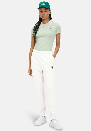 Polo verde chiaro con colletto, pantaloni jogger bianchi e cappellino verde con logo. Sneakers bianche completano l'outfit.