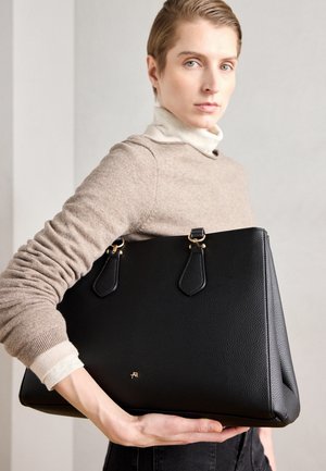 Persona con suéter beige sostiene un gran bolso de cuero negro con herrajes dorados sobre un fondo neutro.
