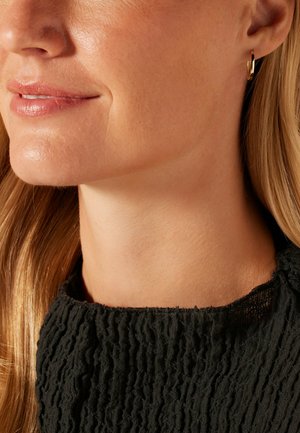 Boucle d'oreille en or avec une surface lisse, portée à l'oreille droite. Le sujet a de longs cheveux raides et un haut noir texturé.