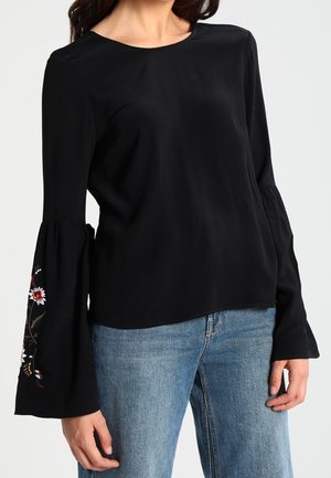 Kvinde iført en sort langærmet bluse med udvidede broderede manchetter og blå jeans, stående med hænderne afslappet ned langs siderne.