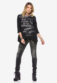 Schwarzer übergroßer Pullover mit weißem Text und abgenutztem Muster, kombiniert mit enganliegenden Jeans aus Denim mit kontrastierenden Nähten und Absatzstiefeln.