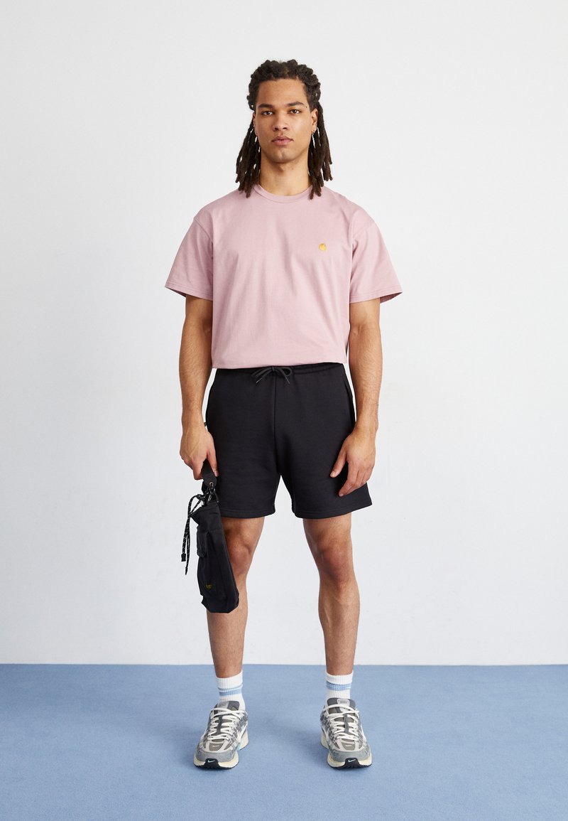 Helles rosa Baumwoll-T-Shirt mit kleinem besticktem Logo, kombiniert mit schwarzen Shorts und grauen Laufschuhen, hält eine schwarze Tasche.