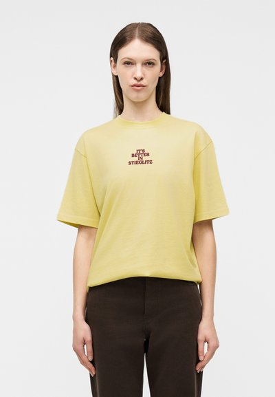 Νεαρό άτομο με μακριά καστανά μαλλιά φοράει κίτρινο T-shirt με κόκκινο κείμενο «IT'S BETTER IN STIEGLITZ» και σκούρο παντελόνι, στέκεται μπροστά σε απλό φόντο.