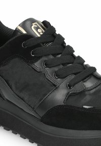 Baskets noires en cuir et en suède, présentant une finition texturée, des lacets ronds et une semelle légèrement compensée. Accent logo doré visible.