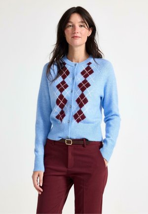 Donna che indossa un cardigan azzurro chiaro con bottoni con motivo argyle color bordeaux e pantaloni bordeaux con cintura nera, in piedi con una mano in tasca.