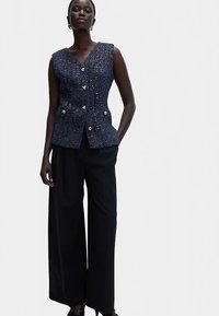 Granatowy bezrękawnik z tweedu z dekoltem w serek, ozdobiony złotymi guzikami i teksturowaną tkaniną, zestawiony z szerokimi czarnymi spodniami.