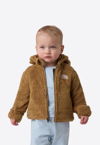 Copil mic purtând o jachetă maro, pufoasă, cu glugă și fermoar, peste un costum albastru deschis, stând pe un fundal alb simplu.