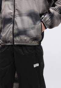 Άτομο που φοράει μαύρο και γκρι ριγέ ζακέτα με φερμουάρ, με το χέρι στην τσέπη, και μαύρο παντελόνι με σήμα λογότυπου EA7 Emporio Armani.