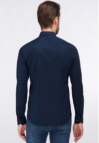 Camisa azul marino de botones con mangas largas, hecha de una tela suave. Presenta un cuello tradicional y puños abotonados. Se muestra la vista trasera.