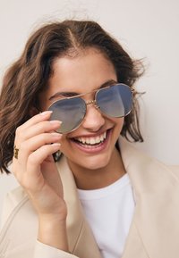 Occhiali da sole aviator con montatura dorata e lenti sfumate blu, caratterizzati da una delicata catena sul ponte, indossati da una persona con una giacca chiara.