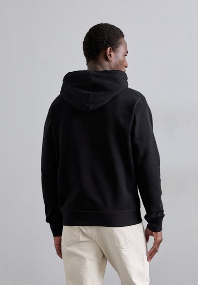 Schwarzer Kapuzenpullover mit lockerer Passform, aus einem glatten Material und mit gerippten Bündchen. Kombiniert mit hellen Hosen.