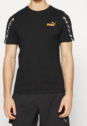 Mann trägt ein schwarzes Puma-T-Shirt mit orangefarbenem Logo auf der Brust und weißem Logomuster entlang der Ärmel, kombiniert mit schwarzen Puma-Shorts.
