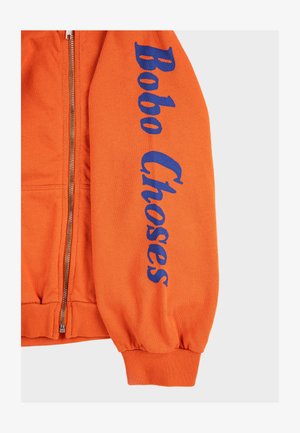 Manica di felpa arancione con zip con testo blu "Bobo Choses" stampato verticalmente lungo il braccio e polsino a coste al polso.