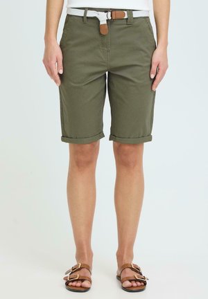 Persoon draagt opgerolde olijfgroene knielange korte broek met wit geweven riem en bruine sandalen met gesp, voor een effen lichte achtergrond.