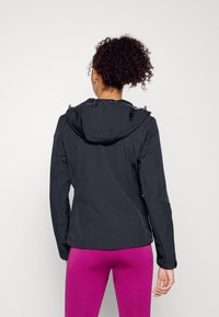 Giacca leggera nera con cappuccio, dotata di zip e polsini elasticizzati. La modella indossa leggings rosa. Texture liscia.
