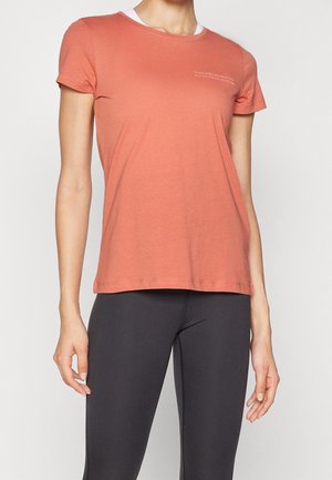 Vrouw die een nauwsluitend koraalkleurig T-shirt met korte mouwen en zwarte legging draagt, staand tegen een effen lichte achtergrond.