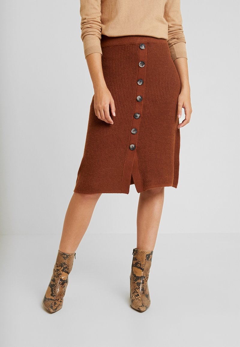 Femme portant une jupe marron côtelée mi-longue à boutons sur le devant, associée à un pull beige et des bottines à motif serpent.