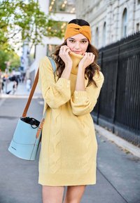 Abito giallo a collo alto lavorato a maglia con motivi a treccia e maniche con bottoni. Il modello tiene una borsa a secchiello azzurro chiaro con una tracolla marrone.