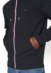 Sudadera negra con cremallera, detalle de cremallera a rayas rojas y blancas, con un bolsillo frontal y acentos de cuero marrón en los puños.