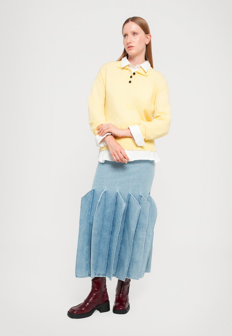 Gele gebreide trui met zwarte knopen, gelaagd over een wit shirt. Lichtblauwe denim rok met plooien en bordeauxrode enkellaarsjes.