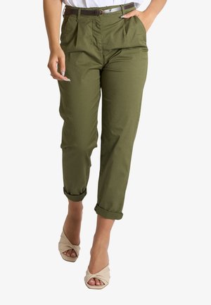 Persona che indossa pantaloni verde oliva risvoltati con cintura marrone, top bianco e tacchi beige aperti attorcigliati, in piedi con le mani rilassate.