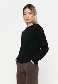 Svart stickad tröja med rund halsringning och långa ärmar, bärs tillsammans med bruna kordflöjlsbyxor. Slät textur, enkel design.