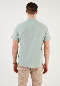 Cămașă scurtă, verde deschis, dintr-un material texturat, cu umeri îmbinați și tiv rotunjit. Afișată cu pantaloni bej; tatuaje vizibile pe brațe.