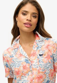 Koszula z krótkim rękawem zapinana na guziki, ozdobiona kwiatowym wzorem w odcieniach pomarańczu i niebieskiego na kremowym tle, z naszywką z logo i kołnierzykiem.