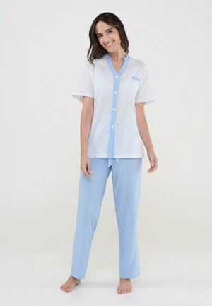 Joven sonriente, con blusa de pijama blanca de manga corta y botones, con lunares azules, y pantalones de pijama azul a cuadros, descalza.