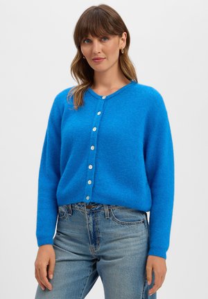 Frau mit hellbraunen Haaren, die einen leuchtend blauen, geknöpften Cardigan und hellblaue Jeans trägt, steht vor einem schlichten weißen Hintergrund.