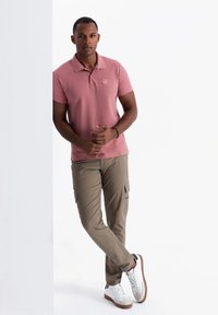 Vaaleanpunainen polo-paita, jossa on pieni logo, khaki cargo-housut ja valkoiset tennarit ruskeilla yksityiskohdilla. Malli nojaa seinää vasten.