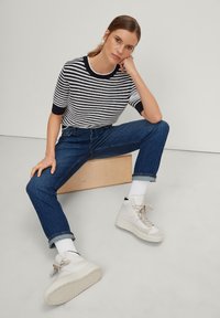 Maglione a righe in navy e bianco, jeans in denim blu e sneaker alte bianche con accenti beige chiaro. Sfondo semplice.