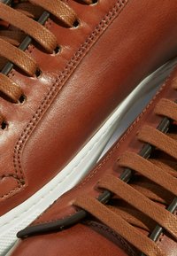 Smeđe kožne tenisice s glatkom površinom, s smešnim vezicama i crnim rupicama za vezivanje. Bijela gumena podloga s teksturiranim detaljima.