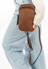 Brown leather crossbody bag with a smooth texture, rectangular shape, zip closure, and adjustable strap, set against light blue denim.