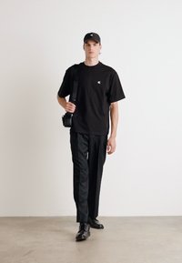 T-shirt nera in cotone con un piccolo logo, pantaloni neri e scarpe lucide nere. Gli accessori includono un cappellino nero e una piccola borsa a tracolla.