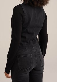 Černá deninová vesta s přiléhavým střihem, kombinovaná s černými elastickými džíny. Mezi prvky patří viditelné švy a dvě nákladní kapsy na zadní straně.