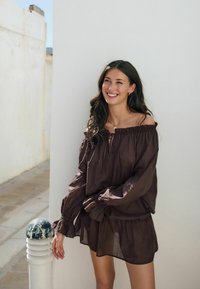 Blusa marrón con escote off-the-shoulder y mangas anchas con puños fruncidos. Tejido ligero, corte relajado, diseño en capas y sutil brillo.