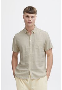 Kortærmet, lys beige linned skjorte med knap-ned krave, enkelt brystlomme og blød tekstur, båret af en person med hænderne i lommerne.