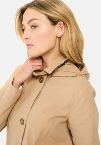 Beige Kapuzenjacke mit strukturiertem Finish, ausgestattet mit zwei großen braunen Knöpfen und einem kontrastierenden Innenfutter. Der Besatz um den Kragen hebt das Design hervor.