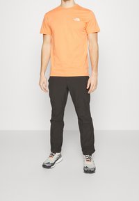 Pomarańczowy bawełniany t-shirt z krótkimi rękawami i białym logo na piersi, zestawiony z czarnymi spodniami i wielokolorowymi butami sportowymi.