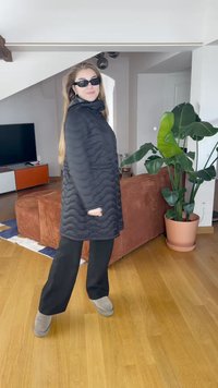 Melna izšūta jaka ar augstu apkakli, vilkta virs melnām platām biksēm. Modelis valkā melnus saulesbrilles un pelēkus potīšu zābakus. Iekštelpu vidē.