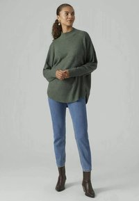 Suéter verde oversized de malha com um decote alto, punhos canelados e fendas laterais, combinado com jeans azuis claros e botas de tornozelo escuras.