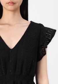 Vestido preto com decote em V, mangas curtas e franzidas, e detalhes em botão. O material apresenta padrões de renda intricados e uma cintura franzida.