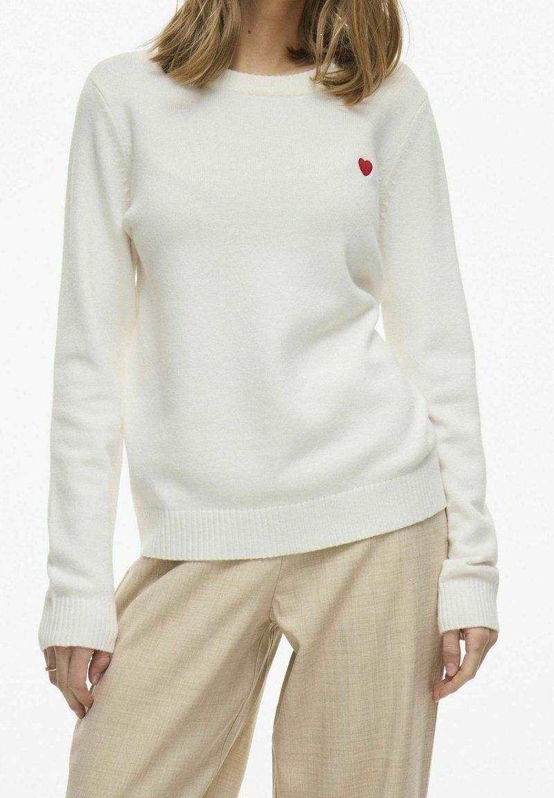 Femme portant un pull blanc à manches longues avec un petit symbole de cœur rouge près de la poitrine, accompagné d'un pantalon beige.